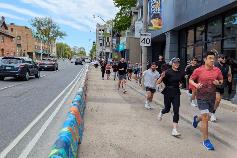 A group of people running on a protected area beside a sidewalk with a busy road on one side and storefronts on the other.